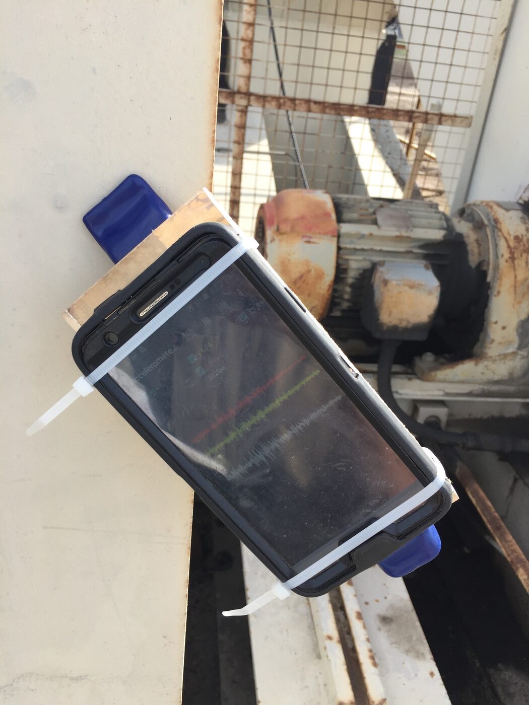 Photo: a smartphone in a rugged case, zip-tied and clamped to a bracket on a rusty pumping-unit motor; its screen shows a three-axis accelerometer app graphing red, green, and blue waveforms.